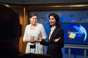 Two women stand in a television studio. One woman is wearing a black blazer and has her hands in an open gesture. There is a television behind them which is showing the text ‘21st Century.’