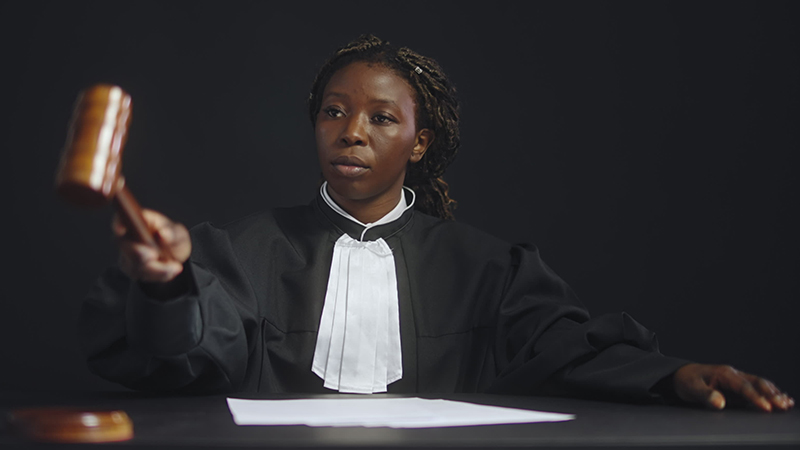 A young African judge wearing her judge's outfit is holding a gavel, about to strike it down on the table in front of her