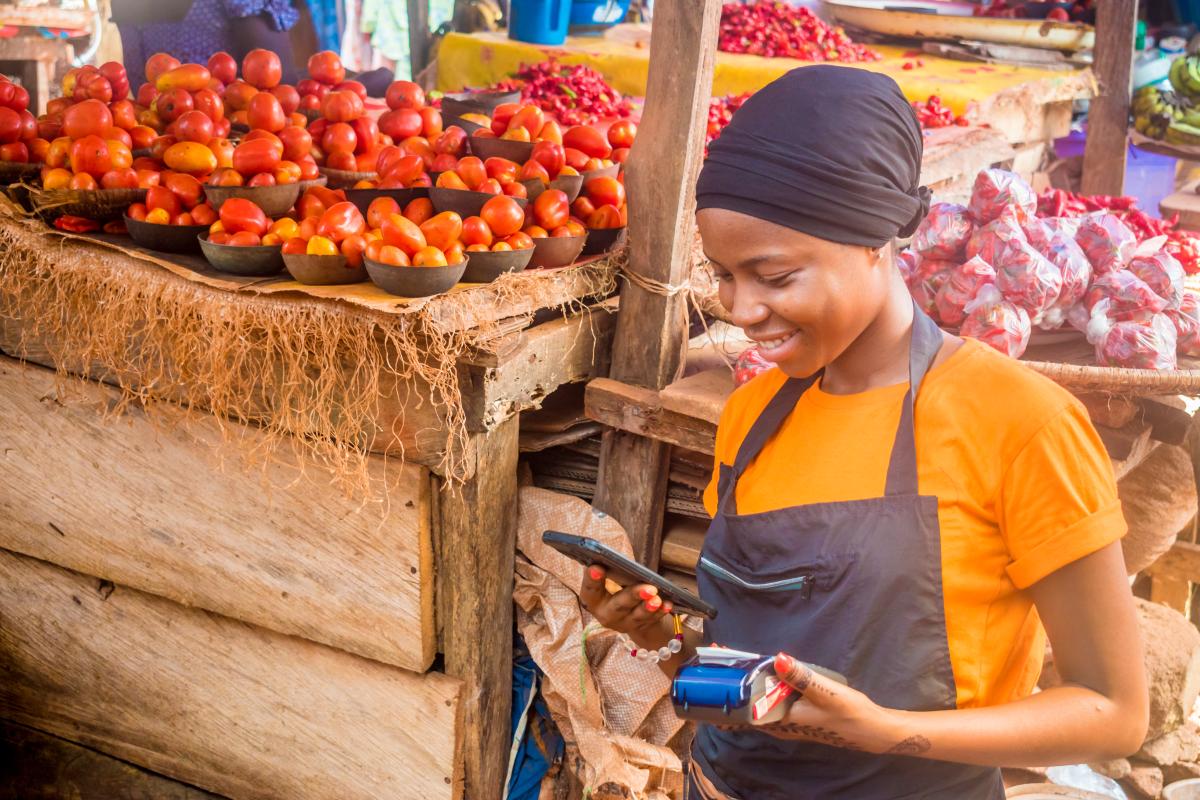 Photo of a woman looking at her mobile phone in front of a tomato vending stand