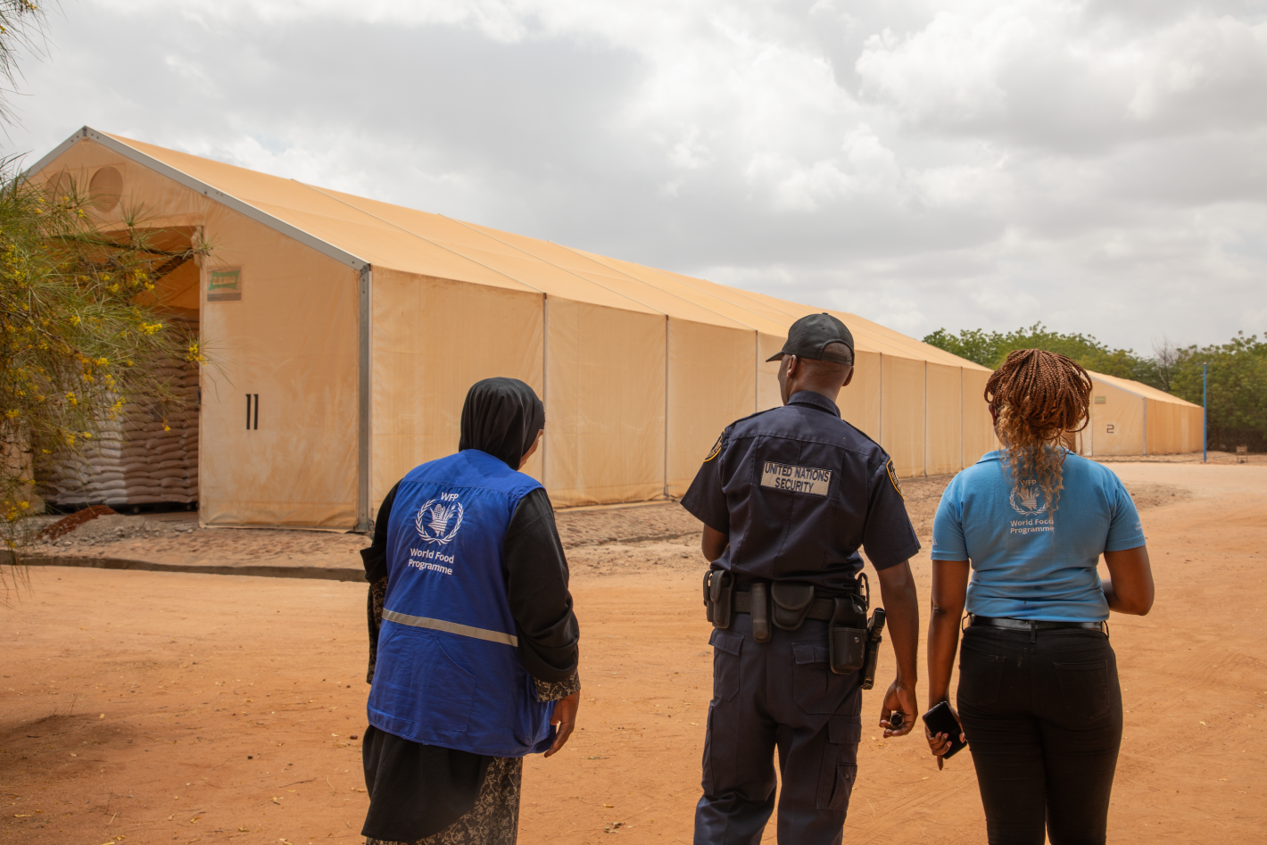 Three people walking on a dusty road with large tents infront of them