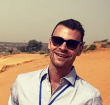 A man wearing sun glasses with a dusty road at the back