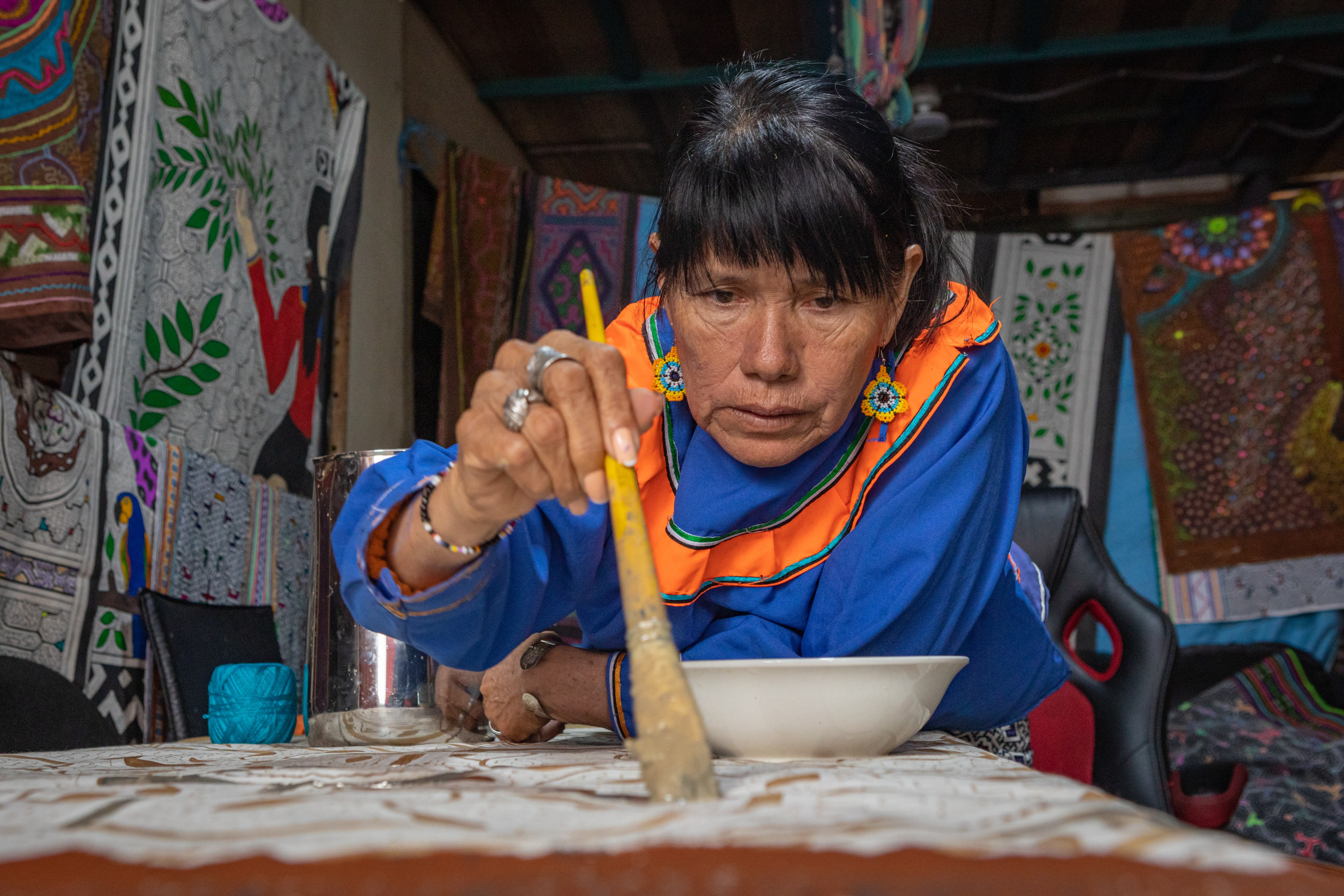 Une femme tient à la main un long pinceau et se penche sur une table de travail pour peindre sur du tissu. 