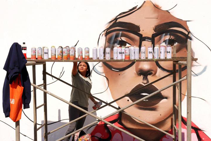 woman with paints in front of graffiti of woman's face