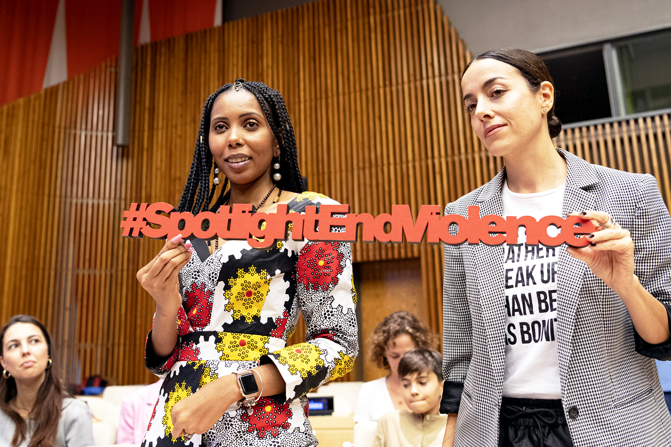 Two women stand up in the GA hall holding a sign that reads: #SpotlightEndViolence.