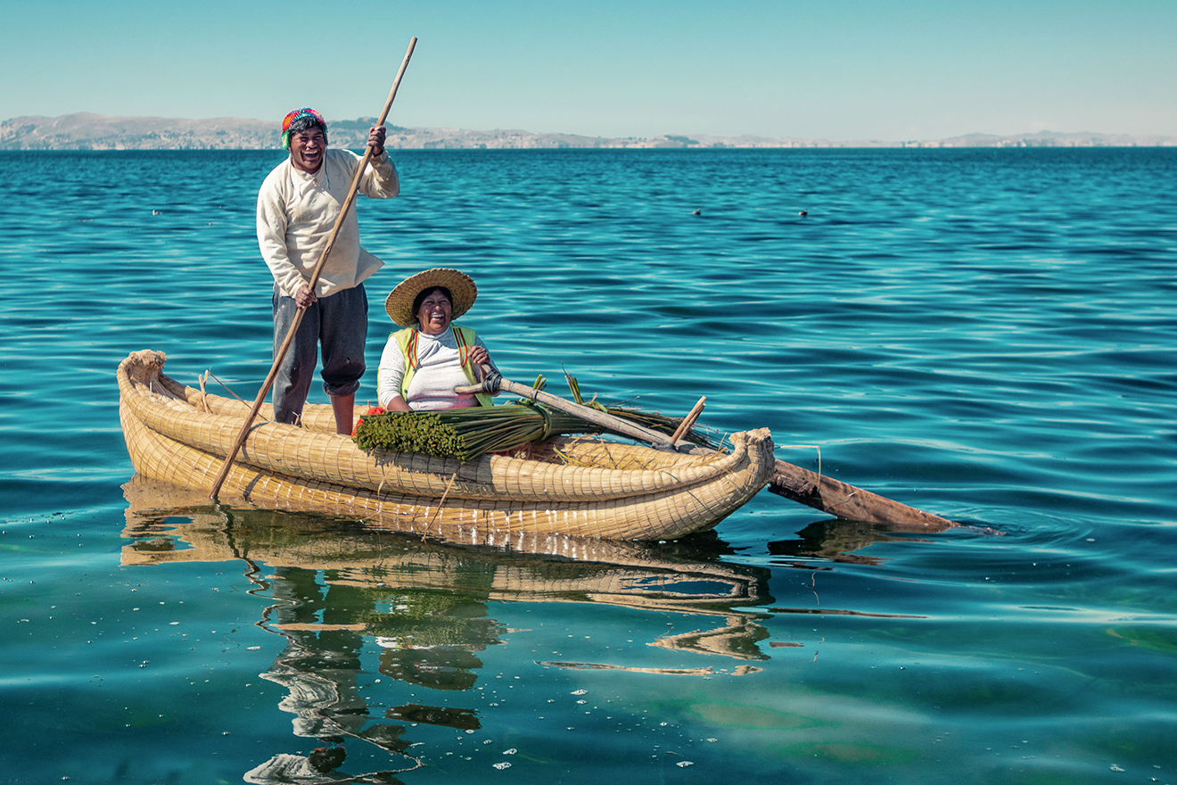 A man stands in a canoe-like boat, paddles it forward, whilst a woman holds a steering oar in place. A bundle of long totora shoots lay across the front of the boat.