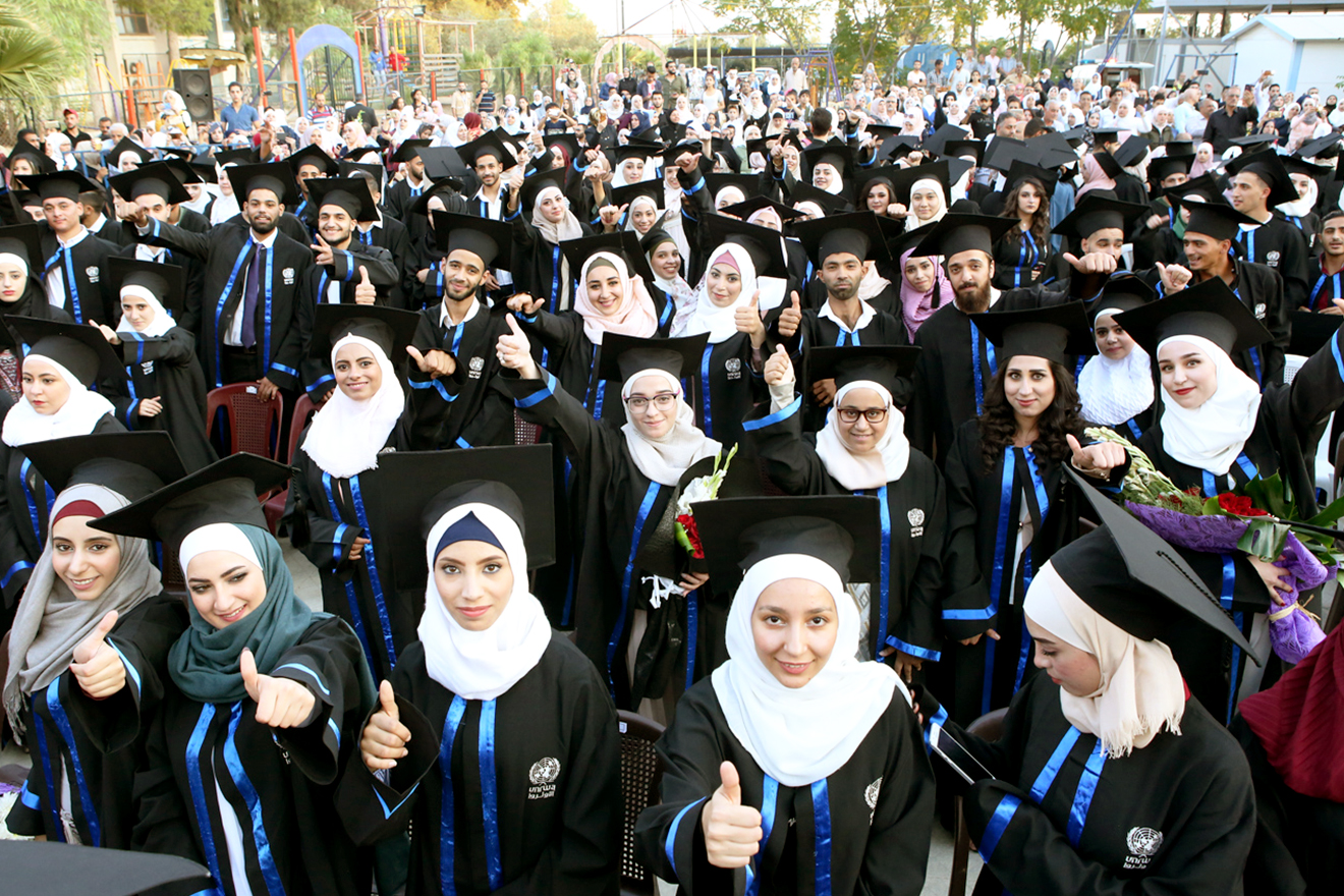 Young women and men in their graduation attire raise a 'thumbs up' as they take a group photo in the playground of a school.