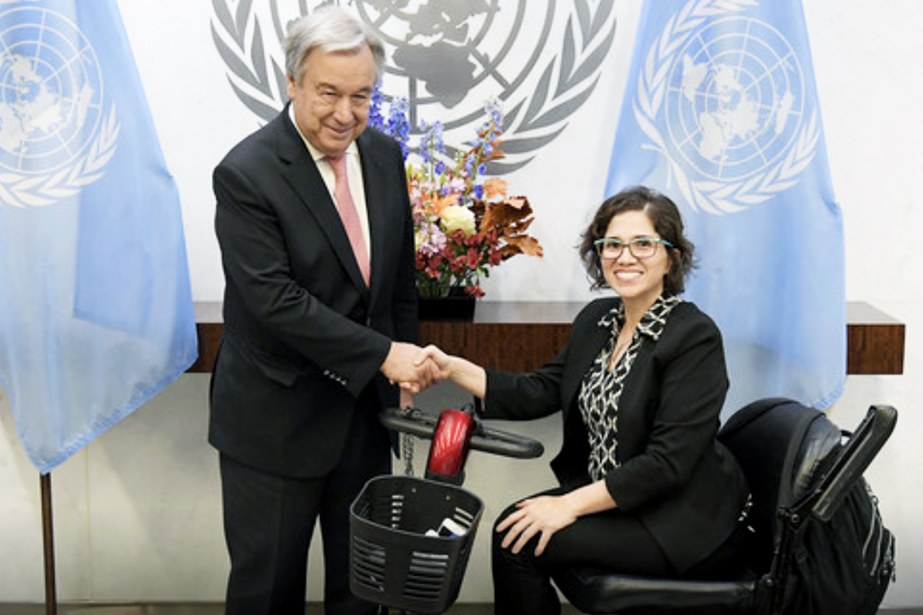 UN Secretary-General stands between two UN flags and shakes hands with Ms. Catalina Devandas Aguilar, who is sitting in a wheelchair.