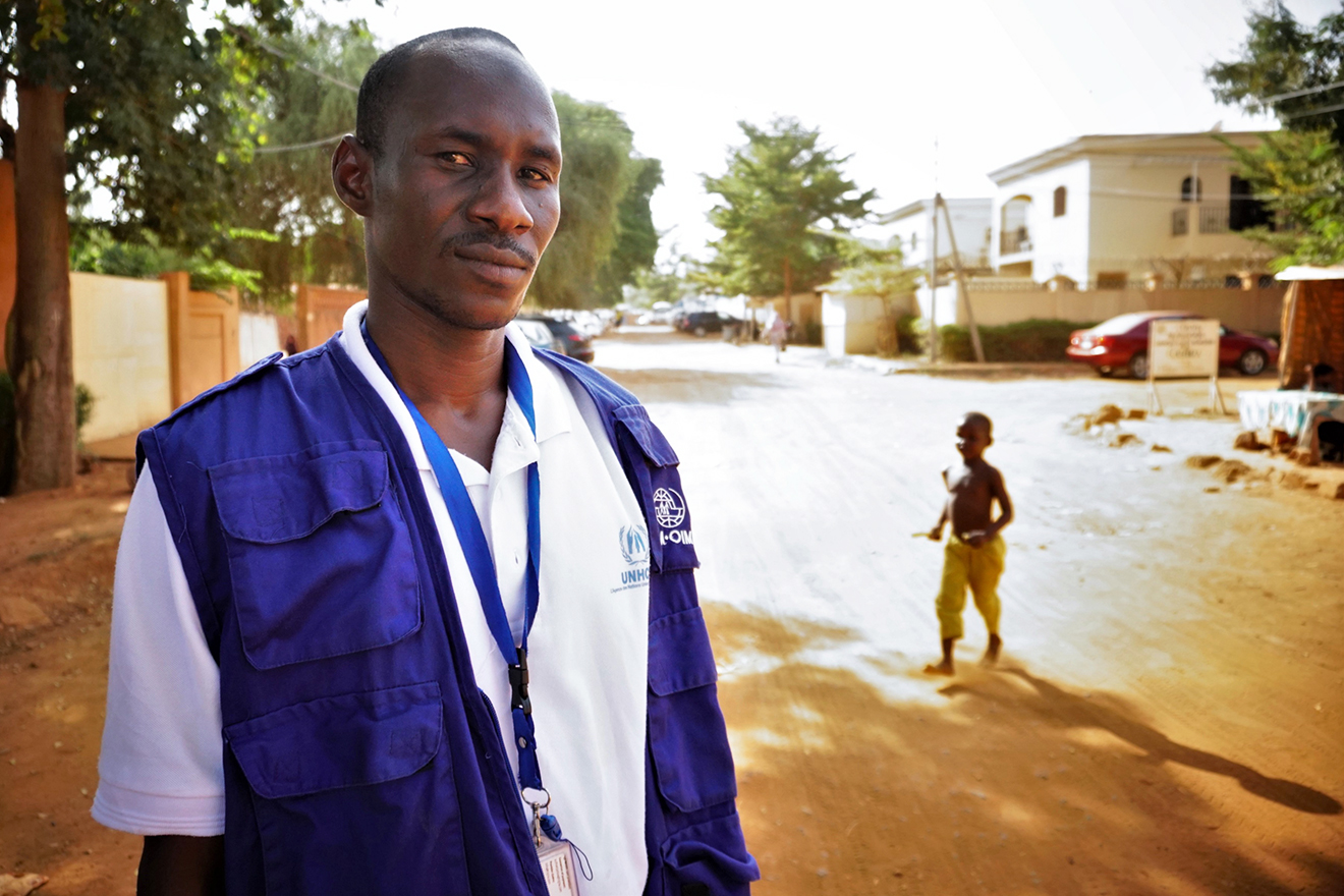 An outdoors portrait shot of Hamma wearing a UNHCR t-shirt and an IOM vest. A child is seen running in the background.