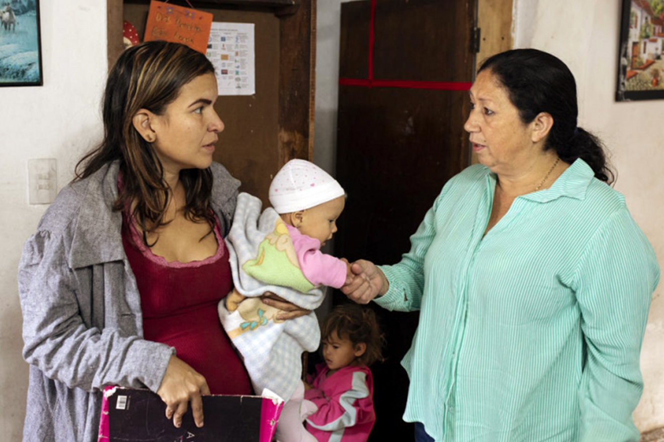 Marta holds a baby girl's hand while talking to the mother who is resting the child on her left hip. A little girl is seen playing behind them.