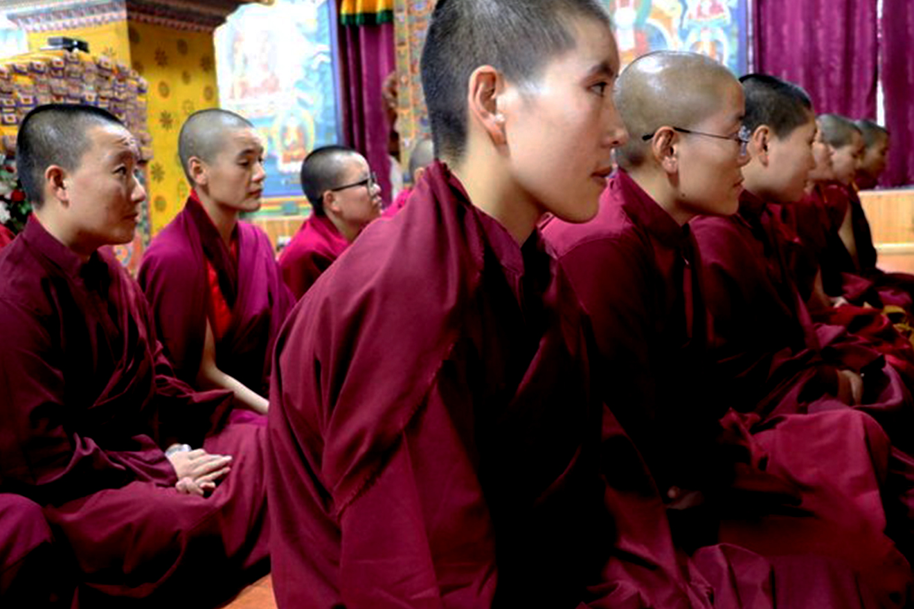 Nuns and monks, with shaved heads and dressed in crimson robes, sit in organized rows as they look ahead in unison.