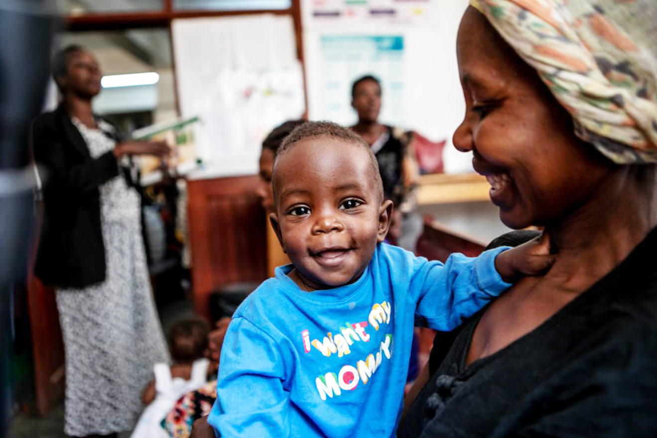 A baby turns to the camera with a big wide grin. The baby wears a t-shirt that says "i want my monny," and is held by the mother.