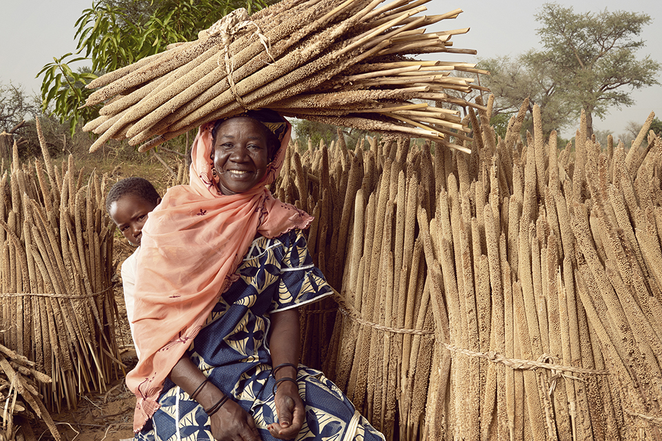 woman carrying bundle of stalks on her head, with child peeking out from behind her