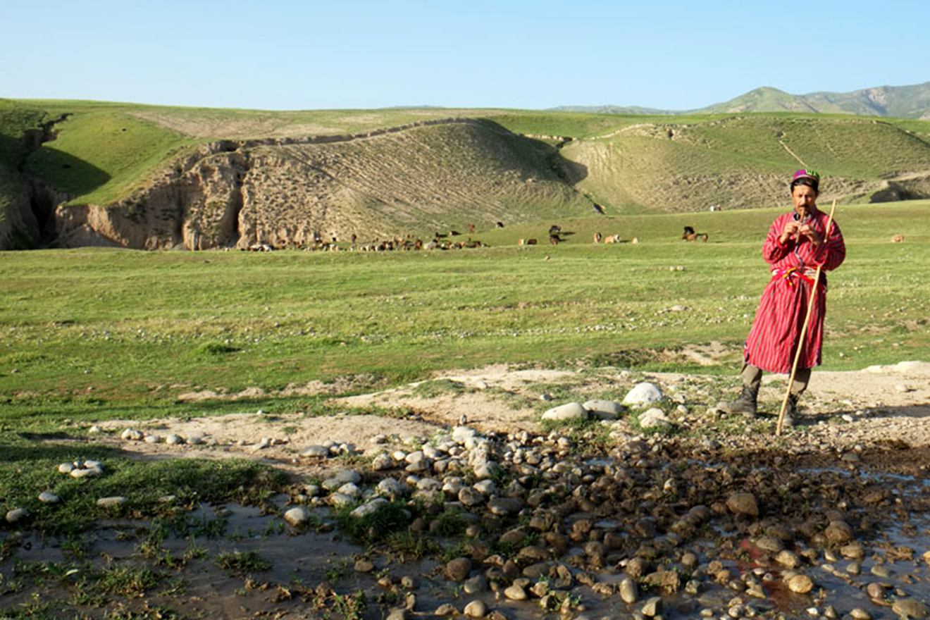 A farmer plays a reed whilst his livestock grazes in the Tajik mountains as seen in the background.