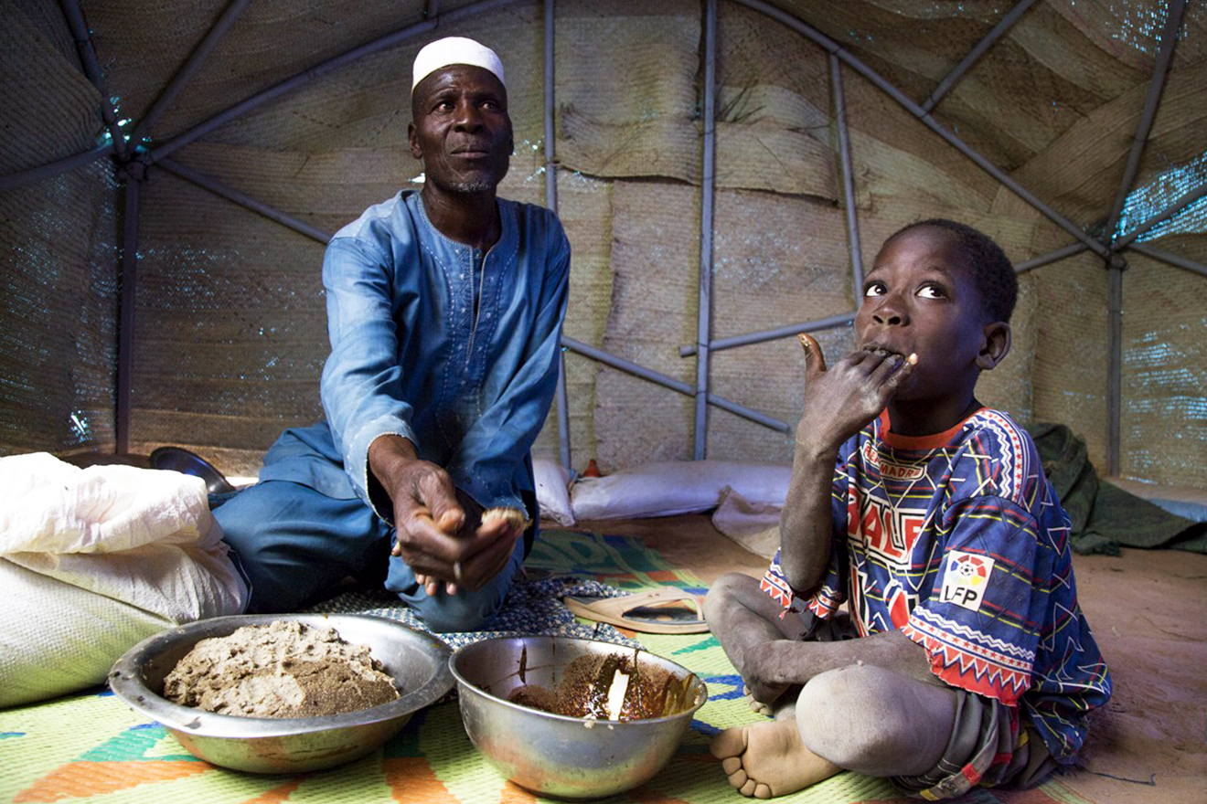 A man and a young boy eat a simple meal together inside a makeshift shelter.