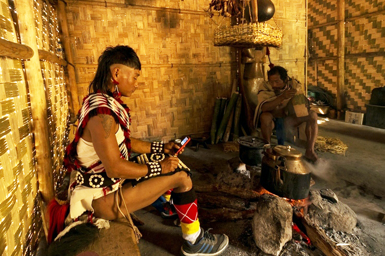 A man from the Nagaland Tribe, wearing indigenous clothes, sits inside a hut made of straw and looks at his smart phone.