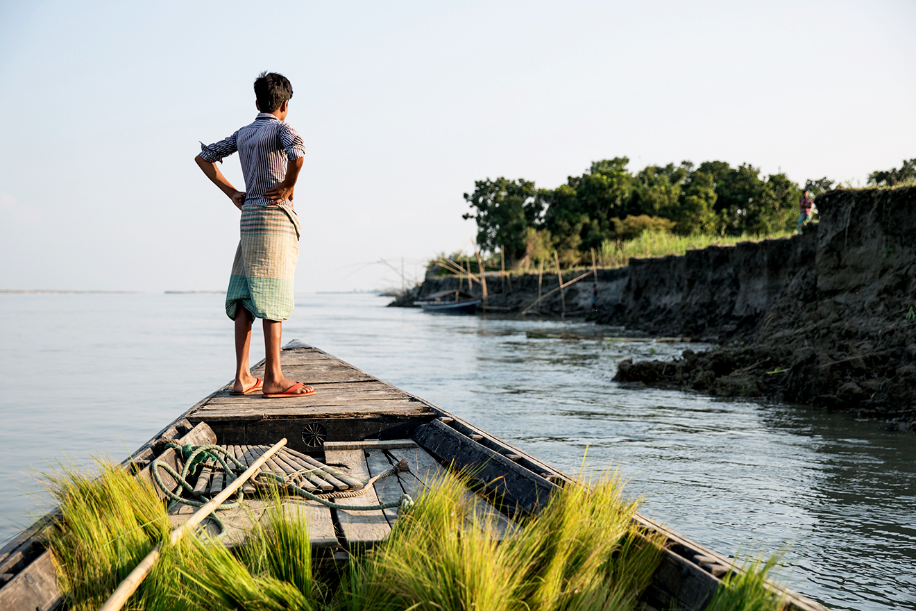 A boy looks out to sea as he stands at the front of a row boat, which has green grass growing inside of it.