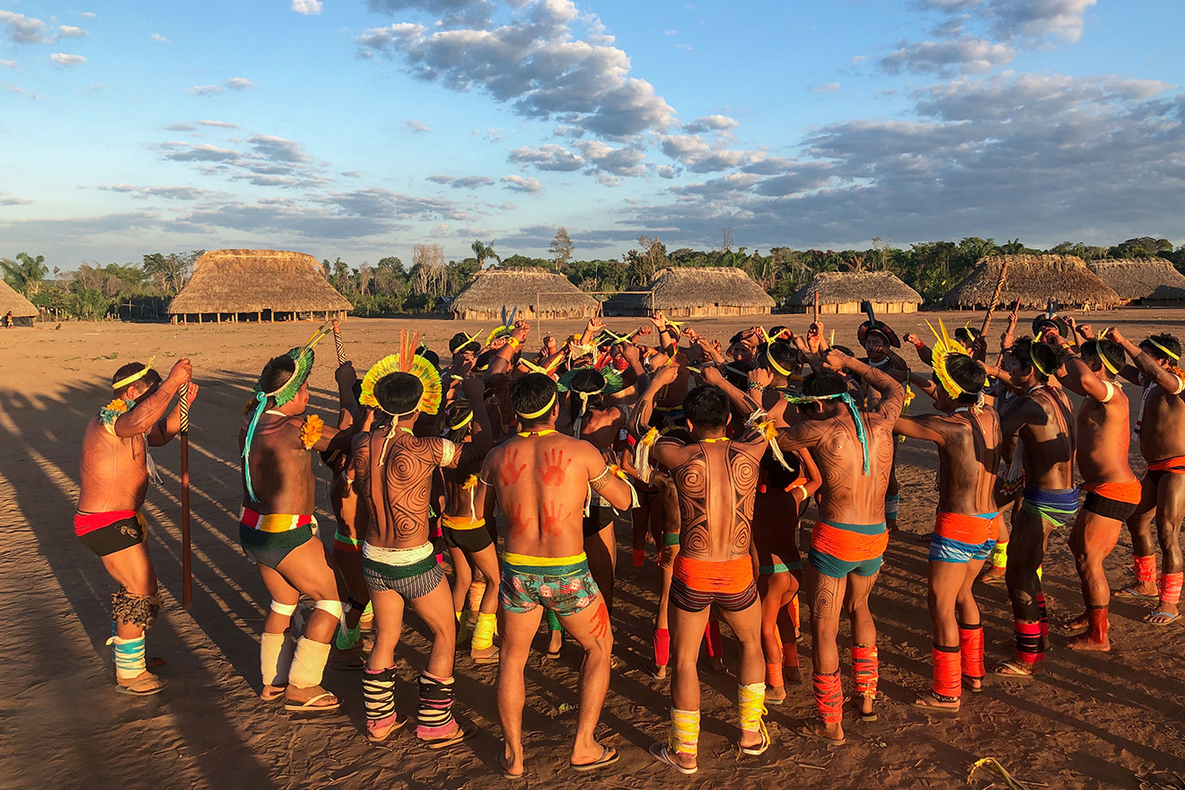 Men dressed in traditional attire gather in a circle in an open space in the midst of traditional homes. They dance whilst the sun is setting, casting a golden glow on the scene.