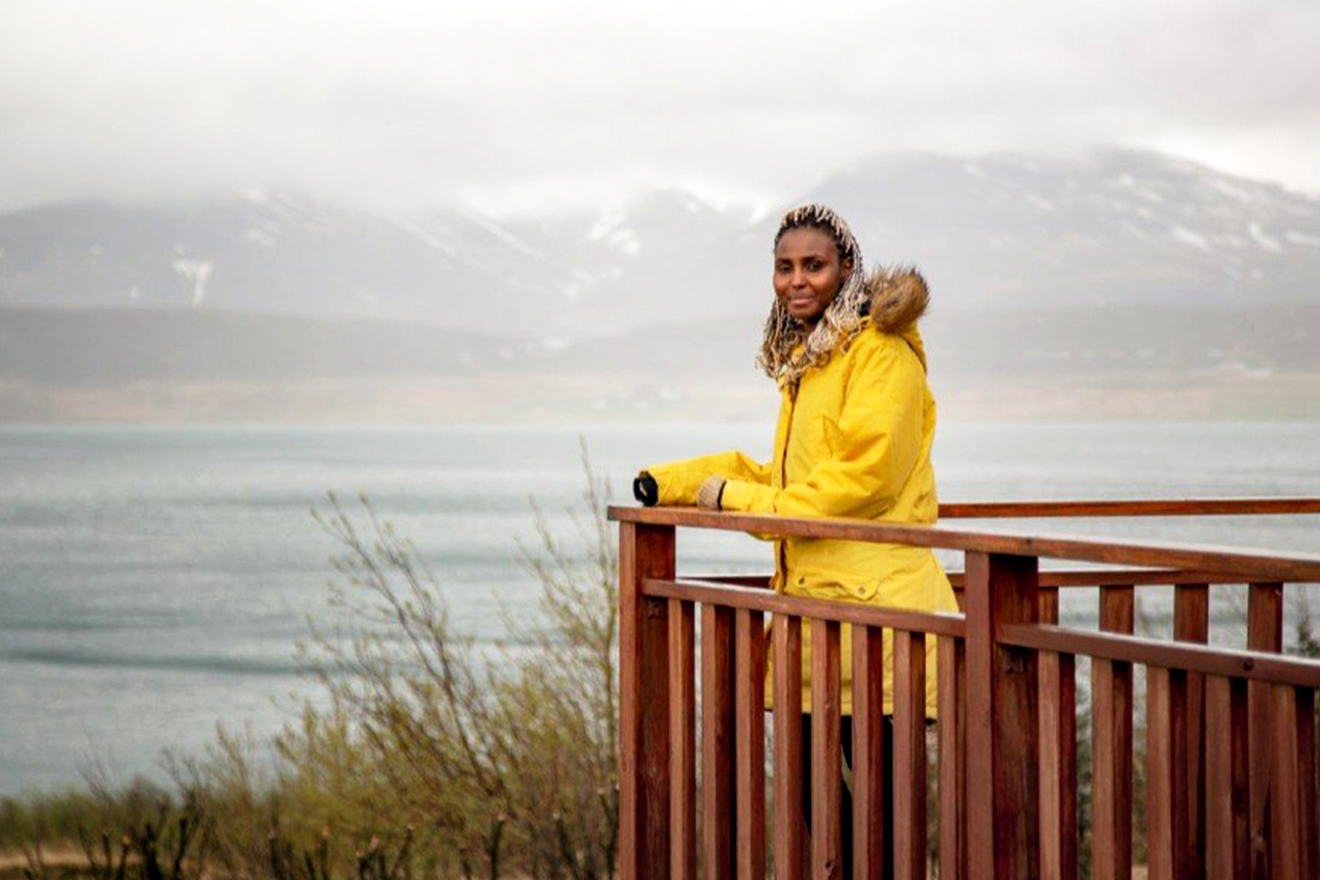 Najmo Fiyasko Finnbogadóttir stands on a deck overlooking a breathtaking view of water and mountains.