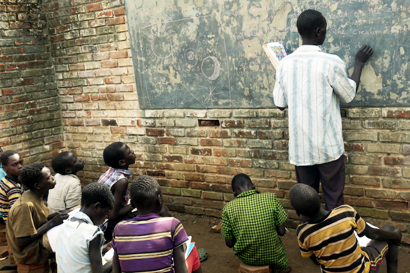 Classroom scene with teacher writing on a chalk-board whilst children sit on stools and look on.