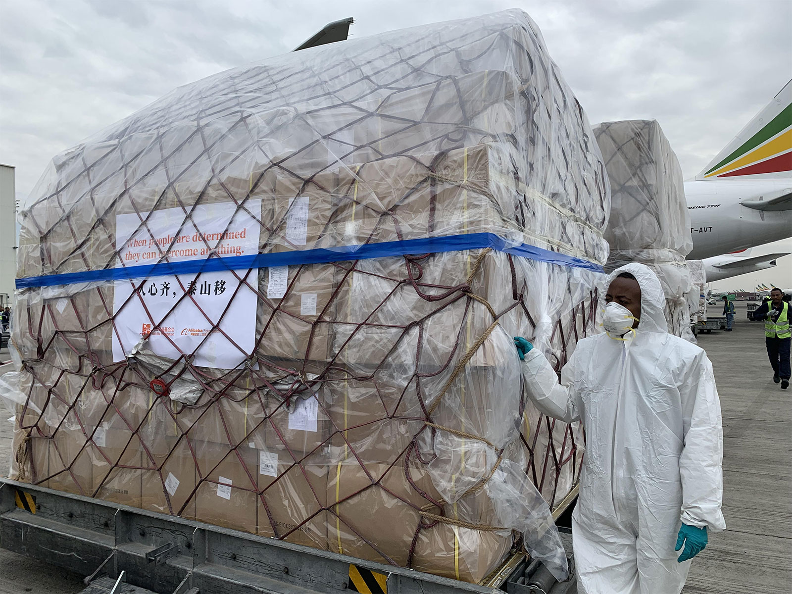 A man wearing tip-to-toe protective gear, including a face mask, stands next to a large shipment in the landing area of aeorplanes.