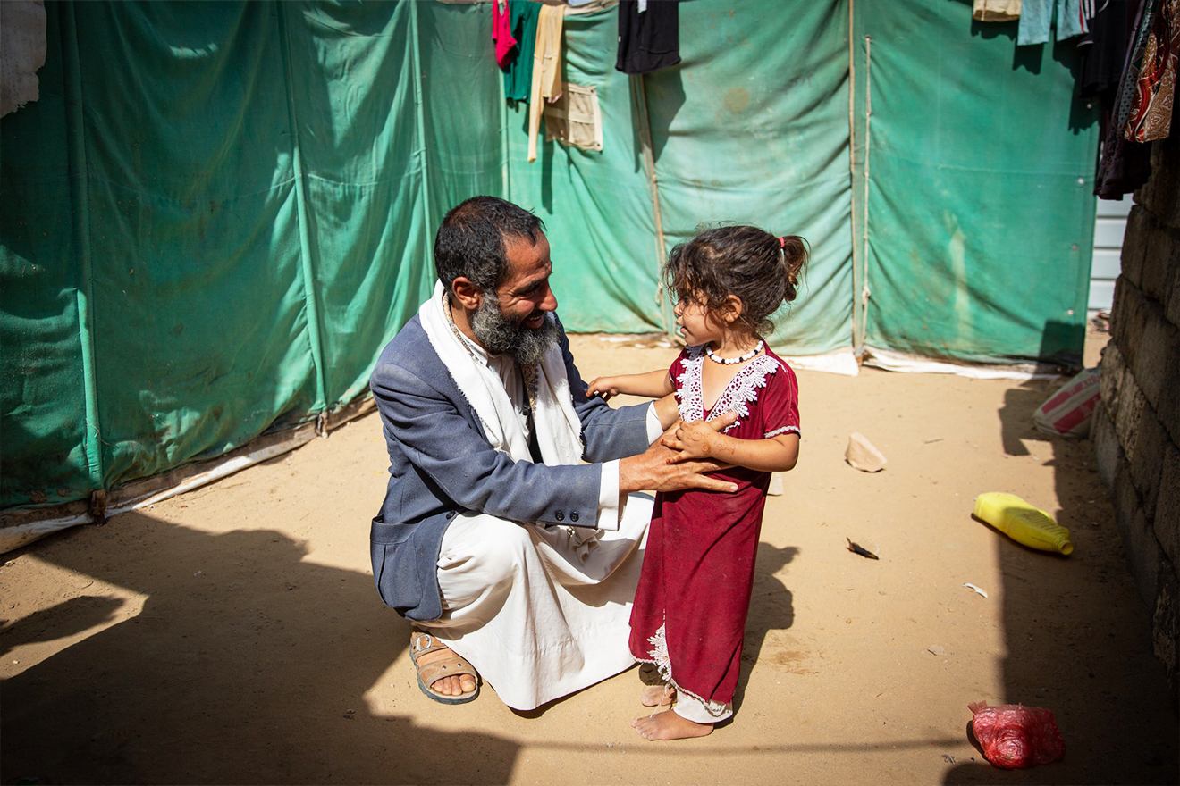 A man crouches to reach the eye level of his daughter and exchanges a warm smile with her.