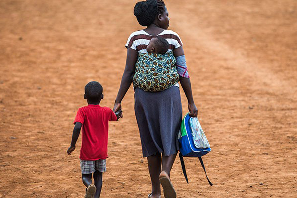 Mother carrying baby on her back and holds son's hand as they walk away on dirt road.