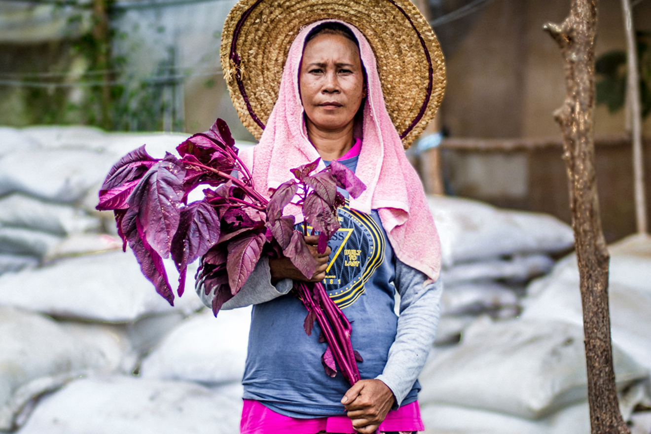 Woman holding a bunch of plants and wearing a straw hat poses for a photo.