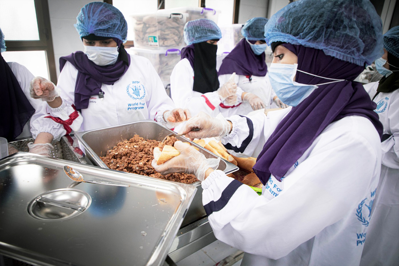 Women wearing masks, white coats and head protection are packing meat.