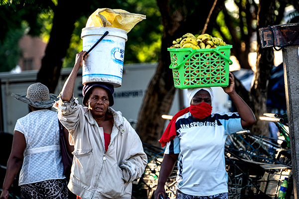 Two women carrying containers and fruit on their heads.