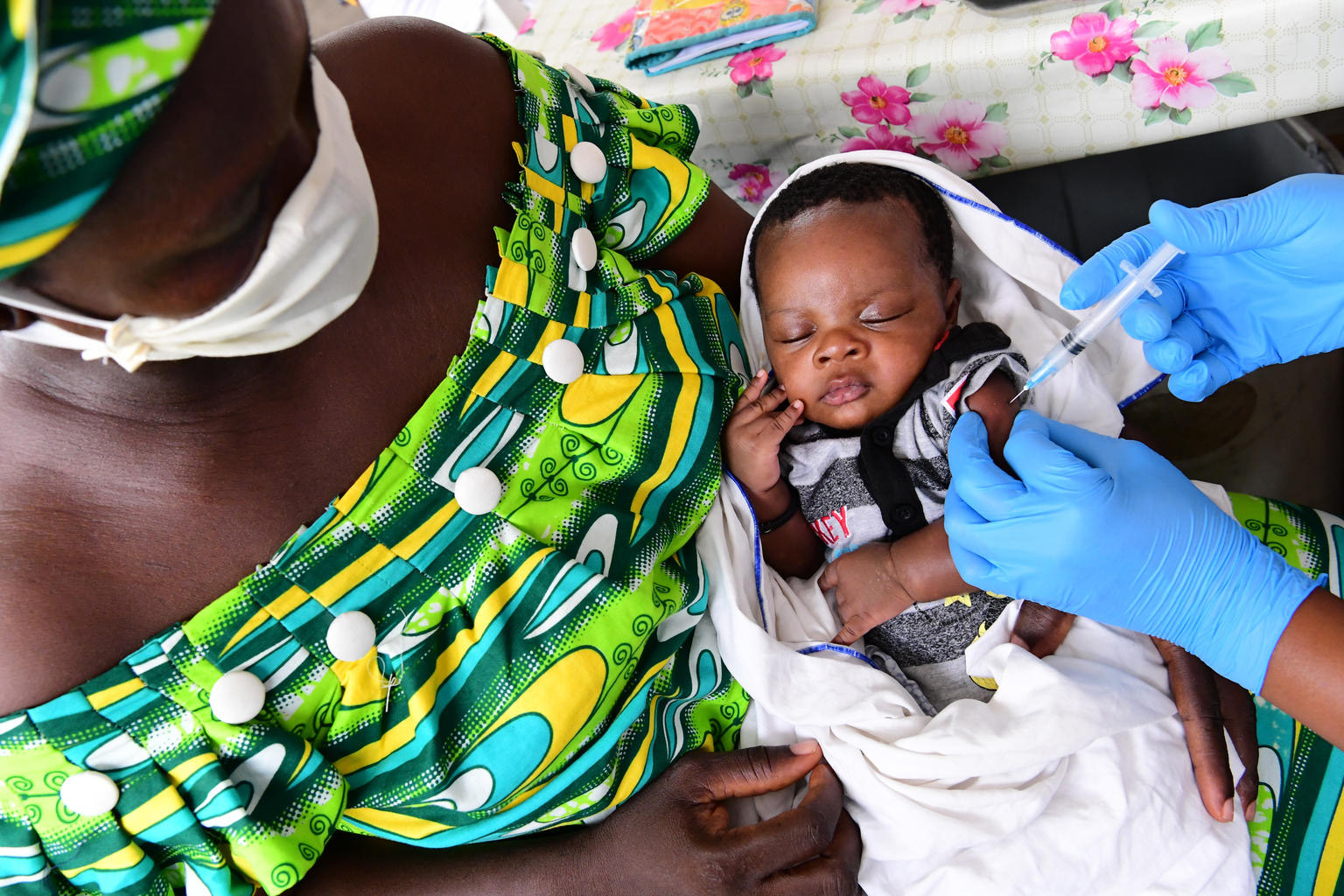 Baby carried by mother wearing a facemask gets vaccinated in the arm by gloved hands. 