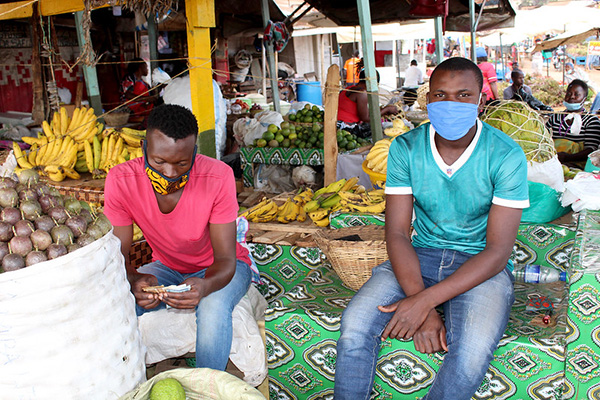 Two men with facemasks sitting side-by-side on a market table.