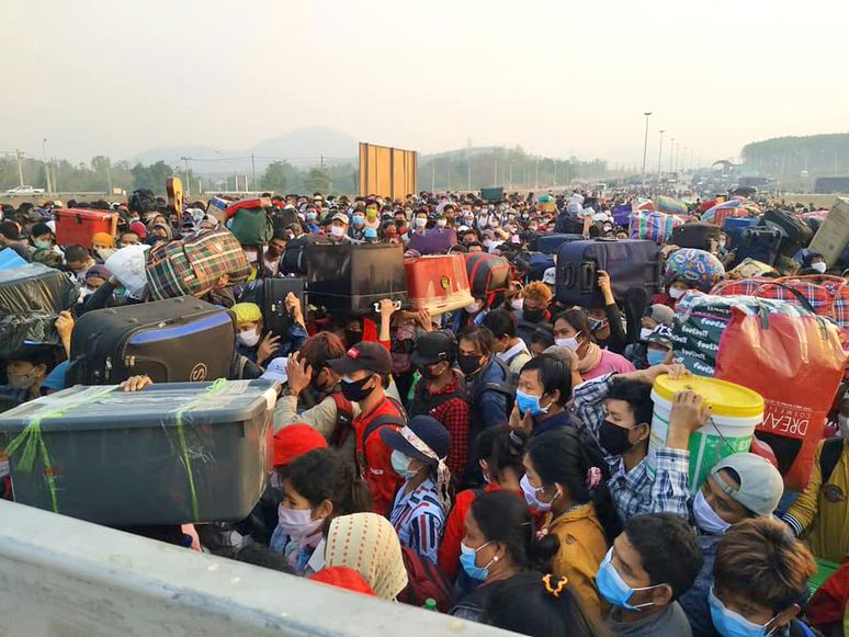 A crowd of people wearing facemasks and carrying luggage stand very closely together. 