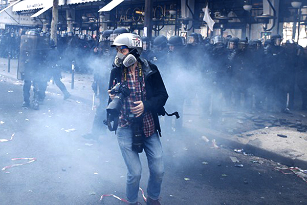 A person wearing a gas mask and holding a camera surrounded by police agents and smoke.