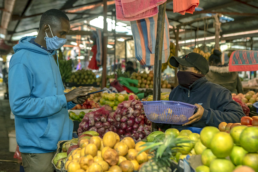Man looks at his mobile phone while a woman waits with a basket of food at the marketplace.