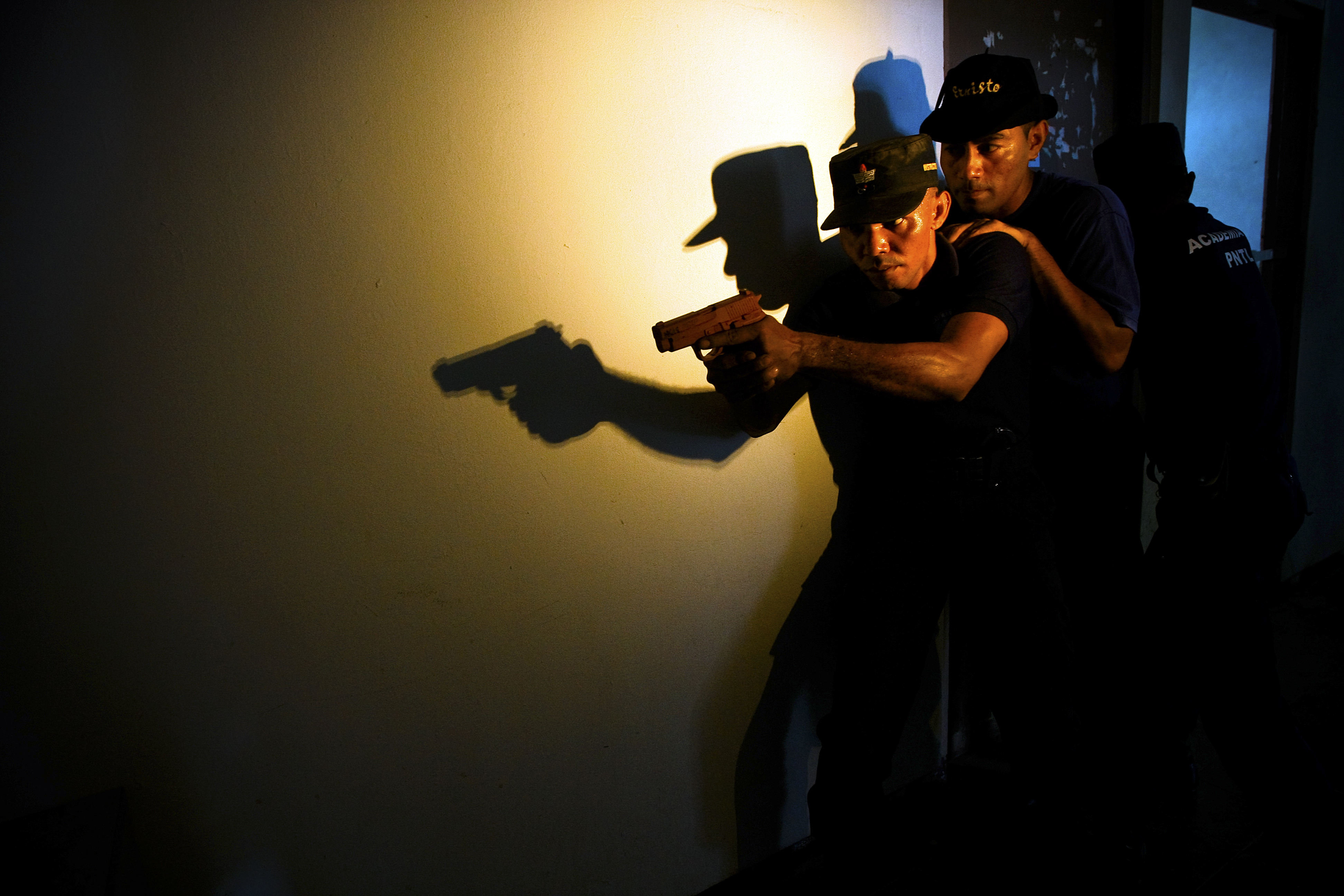 Two security officers are leaning against a wall in a dark interior, one of them holds a gun.