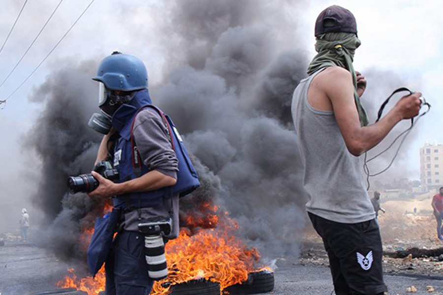 Two men in the middle of a protest with burning tires, one carries many cameras.