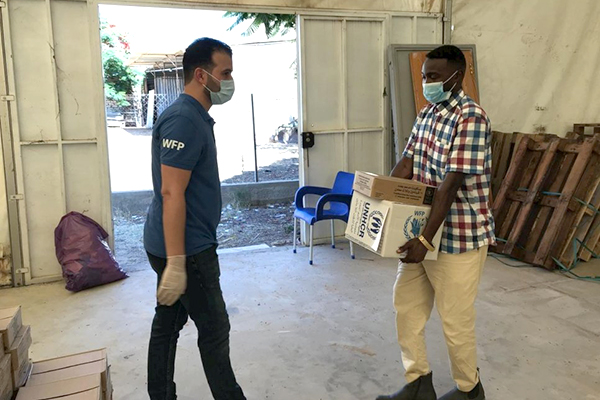 Two men wearing facemasks face each other, one wearing a WFP t-shirt, the other carrying a box labelled UNHCR. 