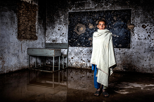 A boy stands in an empty classroom ridden with bullet holes.