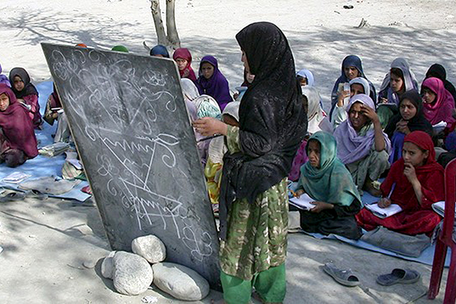 A woman stands outdoors next to a blackboard with girls sitting on the floor facing her. 