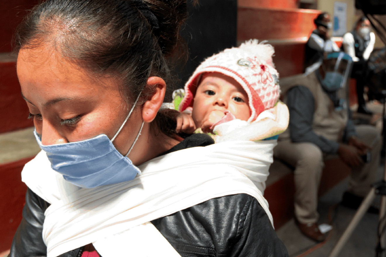 A woman walks through a street wearing a face mask and carrying a child on her back in a sling.