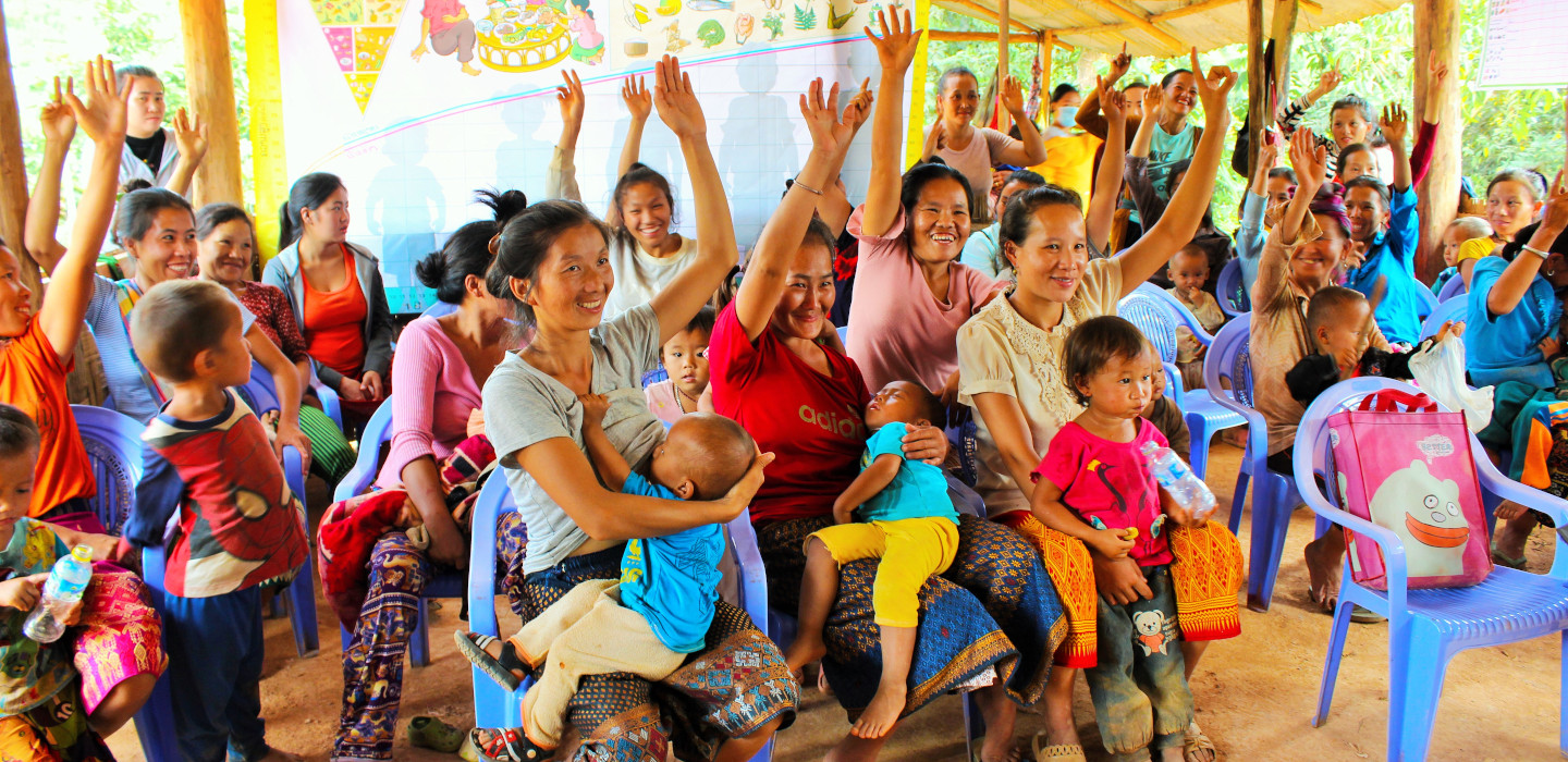 Women sit with children on their laps and raise their hands. 