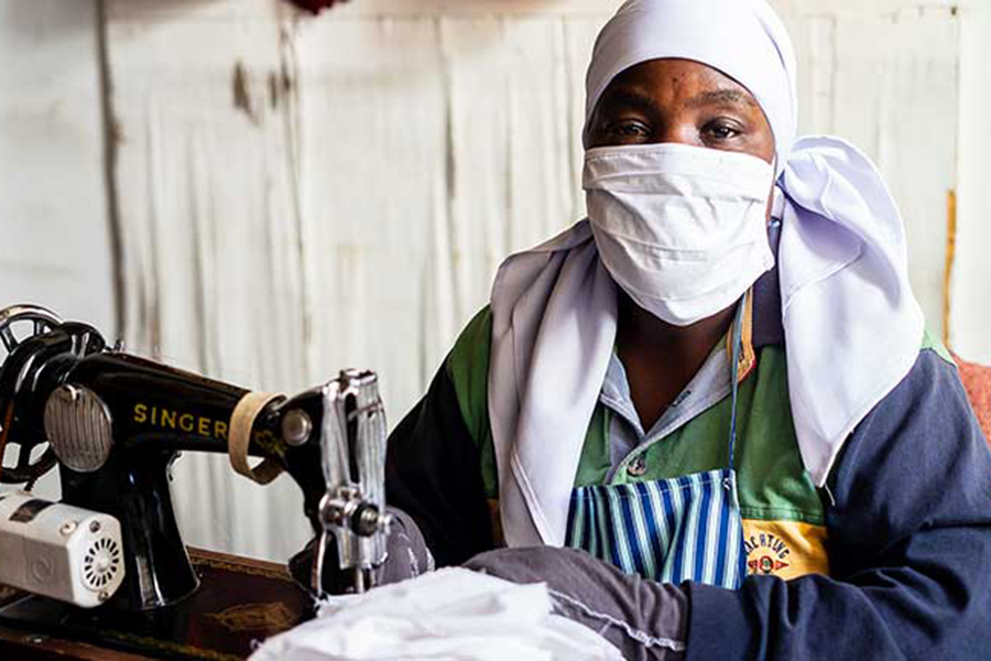 A woman wearing a facemask sits behind a sewing machine. 