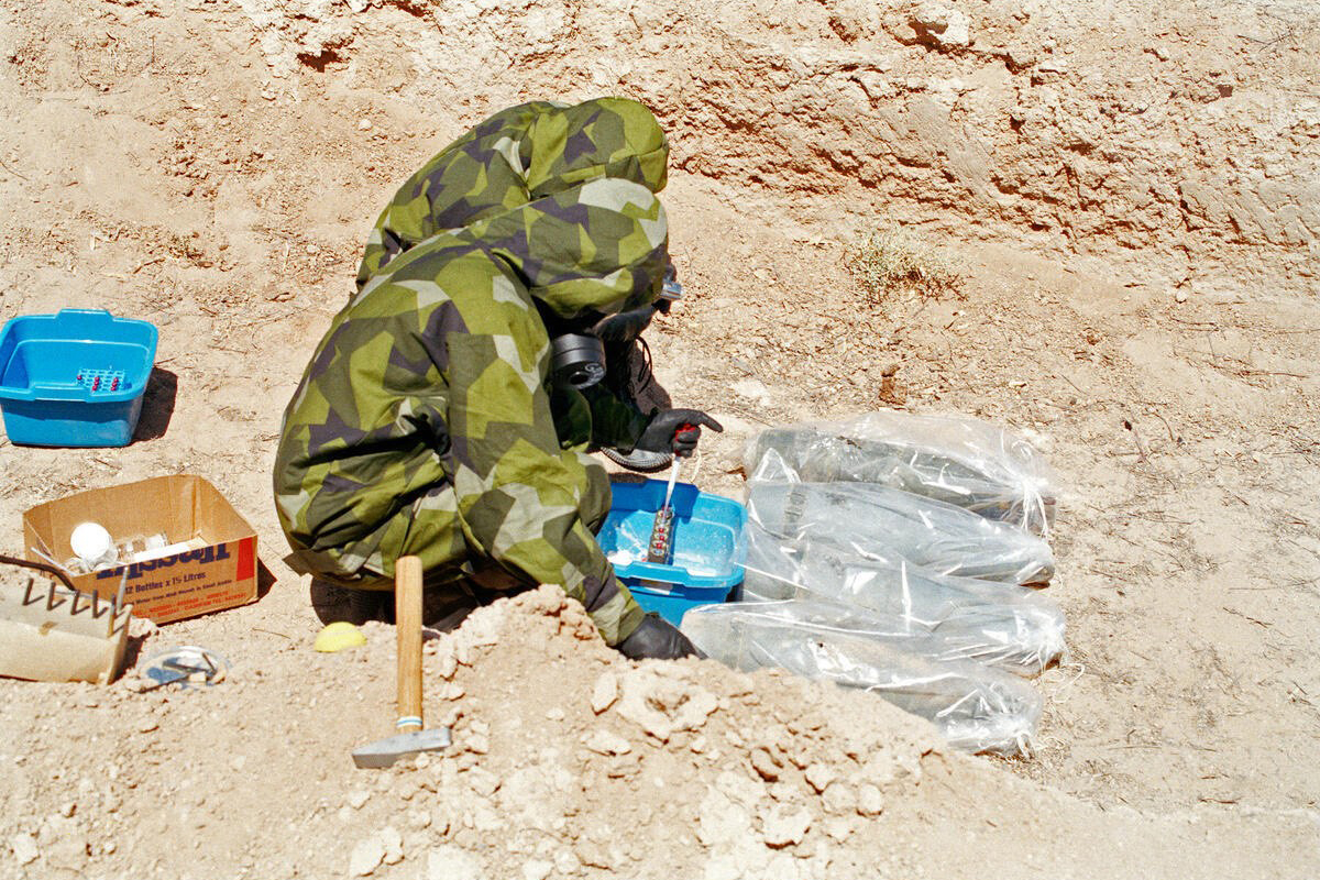 Two people wearing protective suits kneel in front of bagged projectiles in the desert.