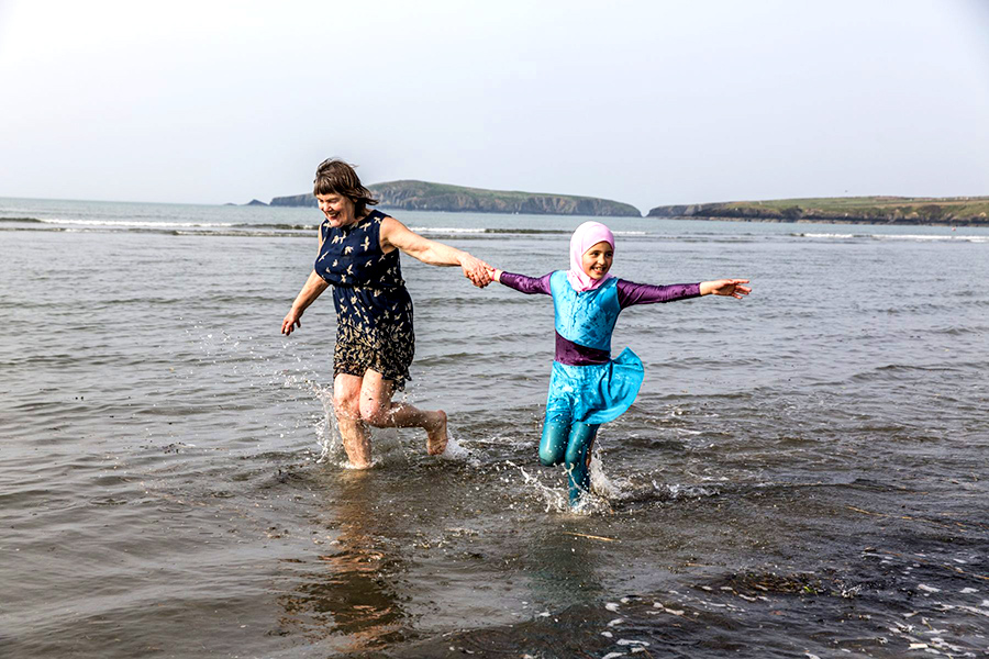 An older woman holds the hand of a girl wearing a hijab while walking on the seashore. 