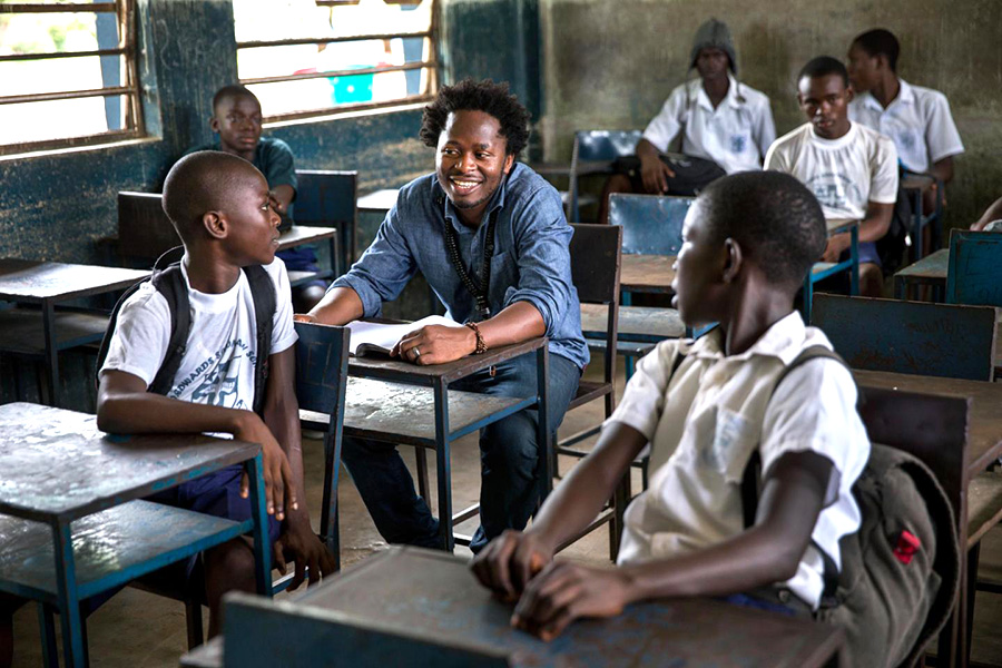 A man sits inside of a school classroom surrounded by children. 