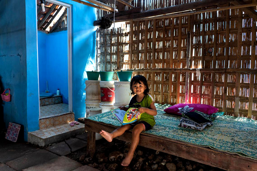 A girl sits on a wooden bed in a room with a latrine next to it. 