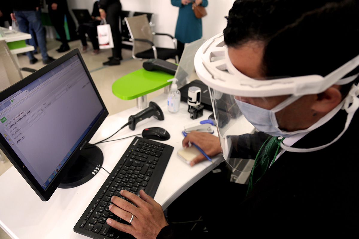 A man sits in front of a computer wearing a mask and face shield.