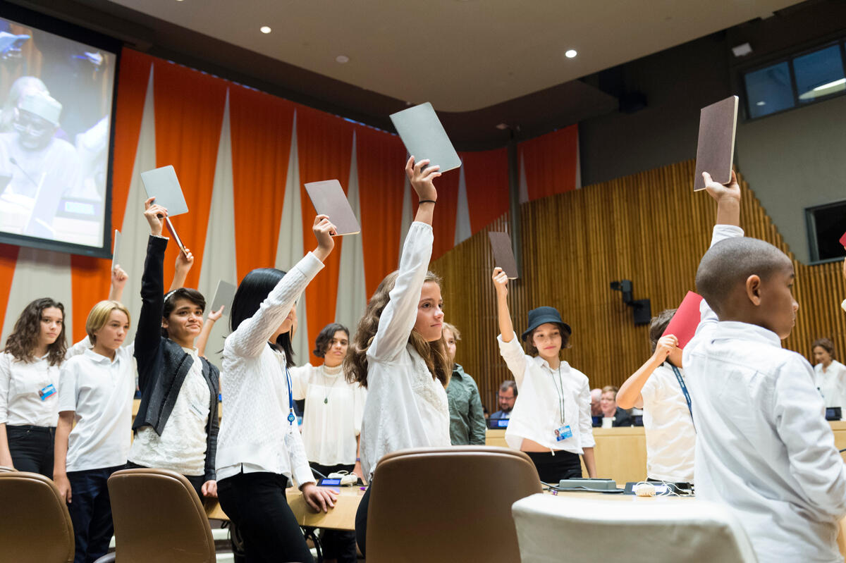 Young delegates stand up and raise their hands holding notebooks in the ECOSOC Chamber in UNHQ, New York.