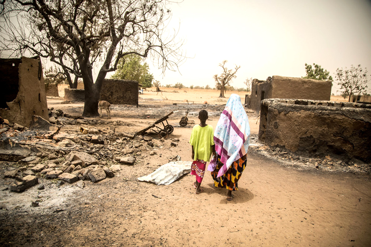 A woman and a boy walk through a destroyed village hand in hand.
