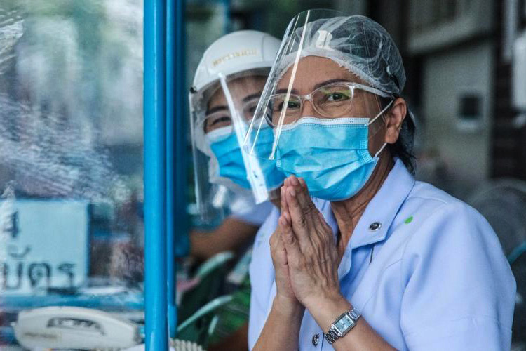 One of two women wearing facemasks and face shields, holds up her hands up together as in prayer. 